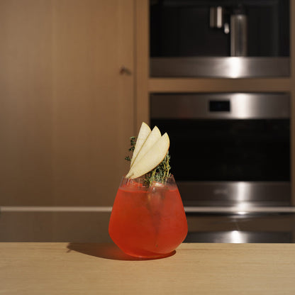 Atelier stemless glass filled with bright red cocktail and garnished with apple and thyme standing on a wooden surface and blurred kitchen appliances in the background
