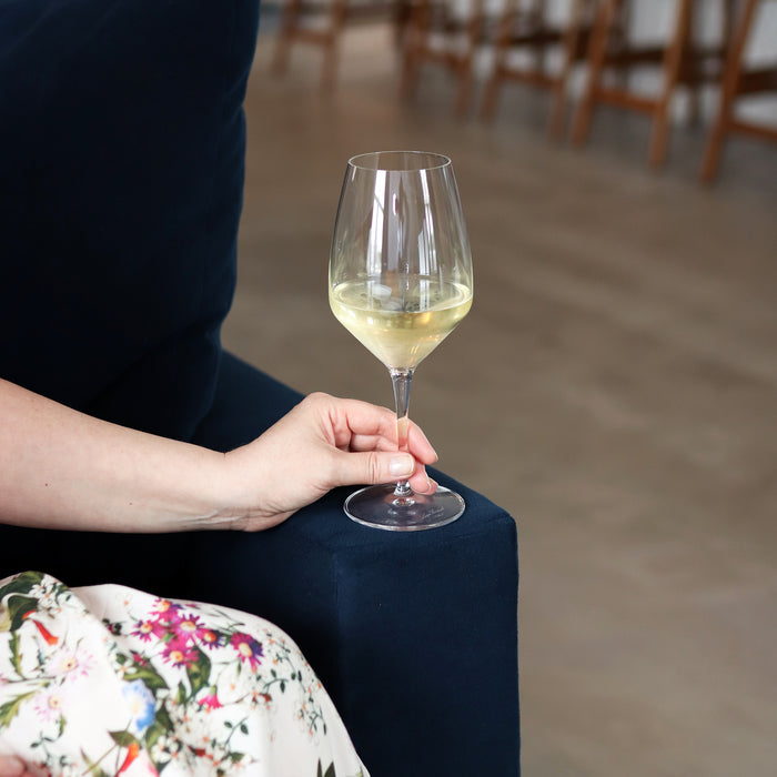Hand holding Atelier Chianti wine glass filled with white wine on a dark blue velvet sofa