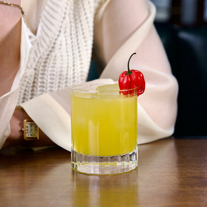 Bach DOF glass with a bright yellow cocktail garnished with a red habanero standing on a wooden surface an a person in ivory outfit in the background