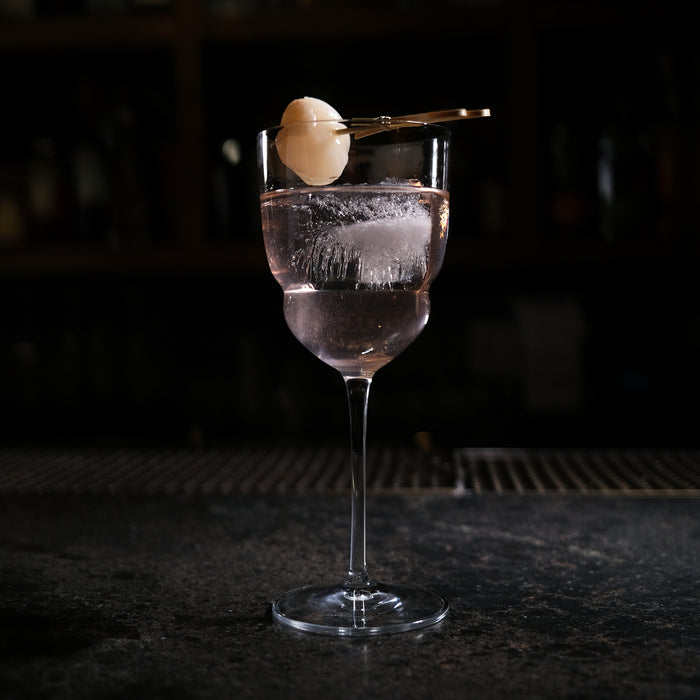 Backdoor '20s Barracuda Cocktail Glass filled with a clear cocktail and garnished with a Lychee on a bar surface and a dark background