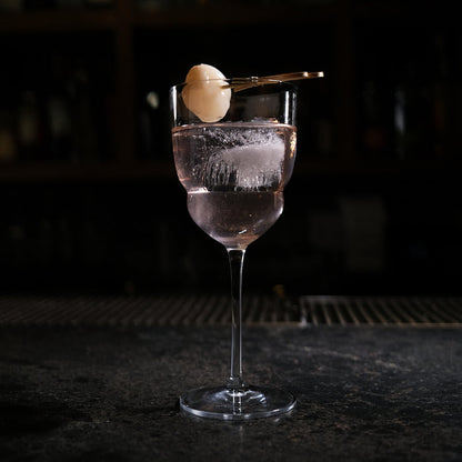 Backdoor '20s Barracuda Cocktail Glass filled with a clear cocktail and garnished with a Lychee on a bar surface and a dark background