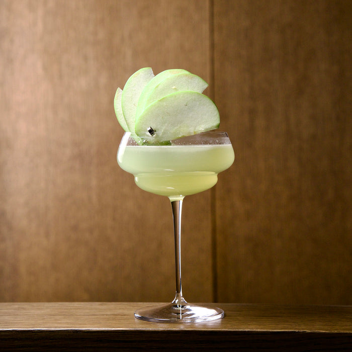 Backdoor '20s Great Gatsby Cocktail Glasses with a light green cocktail garnished with an apple standing on a wooden surface with a wooden wall in the background