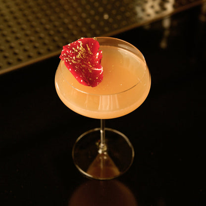 Backdoor '20s Great Gatsby glass with a orange cocktail garnished with a single red rose petal on a dark background
