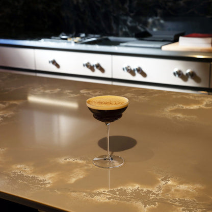 Backdoor '20s Hemmingway Special Cocktail Glass filled with Espresso Martini and garnished with coffee powder, standing on a brown marble surface and blurred kitchen in the background