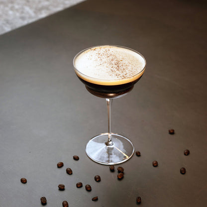 Backdoor '20s Hemmingway Special Cocktail Glass filled with Espresso Martini and garnished with coffee powder, standing on a brown surface decorated with whole coffee beans.
