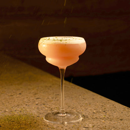 Backdoor '20s Italian Premium Nick & Nora Cocktail glass filled with a pink cocktail and garnished with line zest standing on a marble surface and a dark background