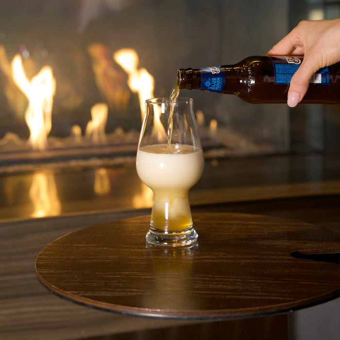 Hand pouring beer from a bottle into a Birrateque IPA Beer Glass standing on a wooden surface and a fireplace in the background