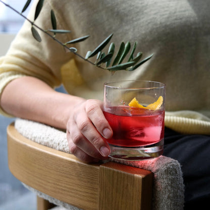 Person holding a glass of non-alcoholic cocktail with a lemon twist and olive branch, sitting on a wooden chair.
