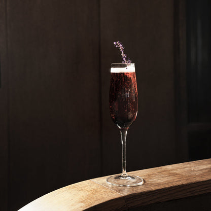Crescendo Flute Glass filled with a deep purple cocktail garnished with lavender standing on a thin wooden surface and a dark background