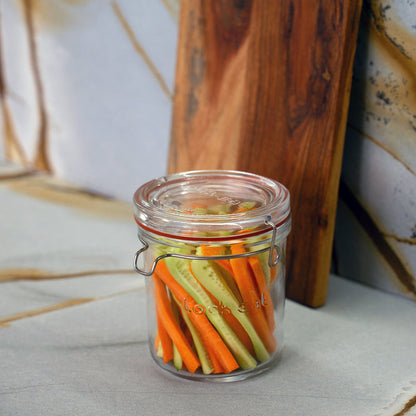 Lock-Eat jar filled with cut cucumbers and carrots standing on a marble surface with a wooden board in the background