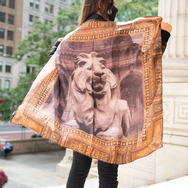 A person holding an oversized silk scarf with an image of a lion and decorative border inspired by library architecture.