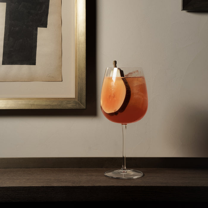 Garnished with a pear slice red cocktail in a Gin & Tonic Jenever glass on a wooden surface with a framed picture in the background