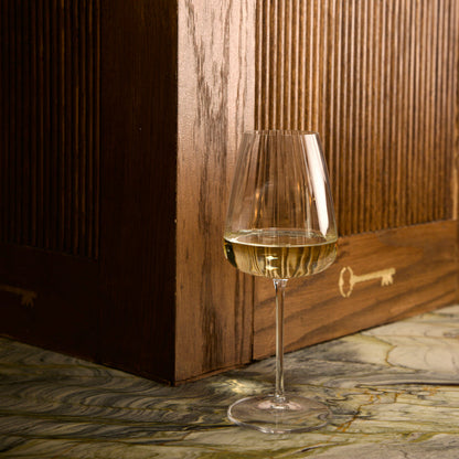 Optica Chardonnay glass filled with white wine standing on a marble surface with a wooden wall background