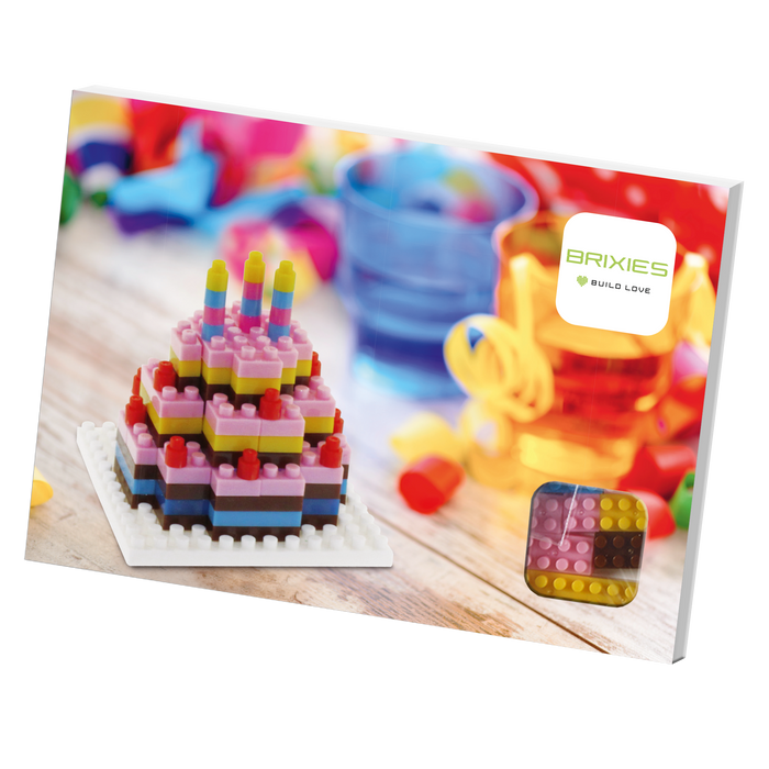 A colorful Brixies Postcard Birthday Cake toy brick model with candles sits on a table among party decor and bright cups. This unique gift is a miniature build kit that can be mailed, just like the Brixies Build Love box in the background.