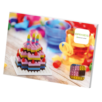 A colorful Brixies Postcard Birthday Cake toy brick model with candles sits on a table among party decor and bright cups. This unique gift is a miniature build kit that can be mailed, just like the Brixies Build Love box in the background.