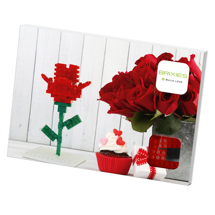A Brixies Postcard Flower, a bouquet of real red roses in a metal vase, a decorated cupcake, and a gift box are displayed on a white surface with a Brixies Build Love sign in the background.