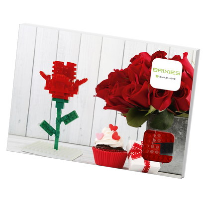 A Brixies Postcard Flower, a bouquet of real red roses in a metal vase, a decorated cupcake, and a gift box are displayed on a white surface with a Brixies Build Love sign in the background.