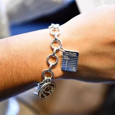 Close up photo of model's wrist with silver charm bracelet and silver library card charm and circular NYPL lion logo charm. Top of library card charm is  imprinted 'The New York Public Library'.  On lines below are imprinted 'Title:' and 'Author:' and the bottom of the charm has NYPL's lion logo.