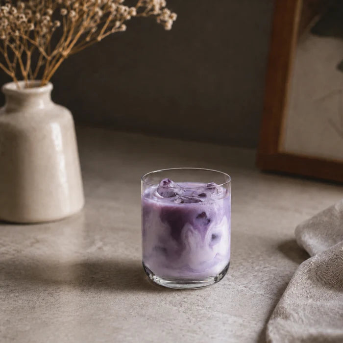 A clear glass tumbler filled with an ube latte, set on a textured stone surface with a ceramic vase and dried flowers in the background.