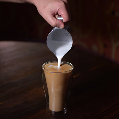 Thermic double-wall latte cup filled with latte and a hand pouring milk inside, standing on a dark wooden surface