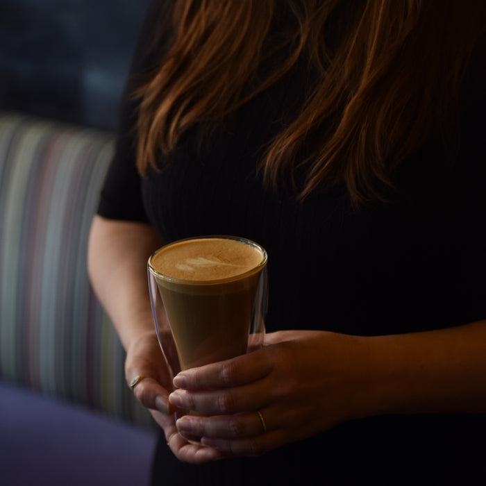 Two hands holding a Thermic double-wall latte cup filled with latte on a dark background