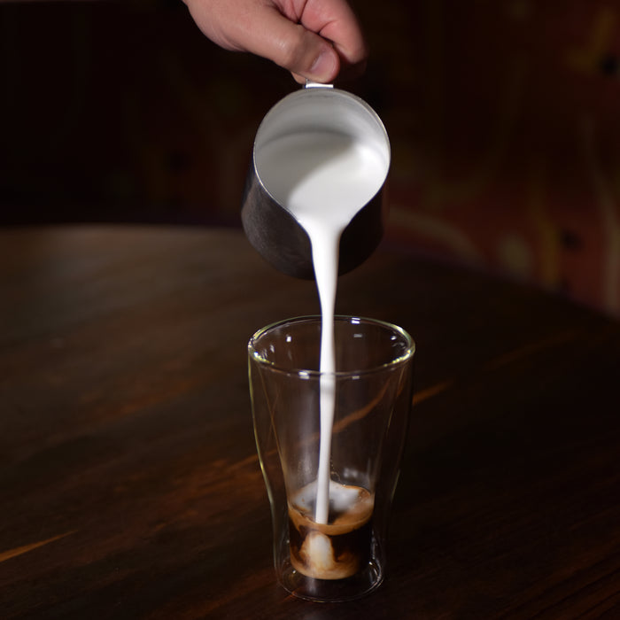 Thermic double-wall latte cup filled with latte and a hand pouring milk inside, standing on a dark wooden surface