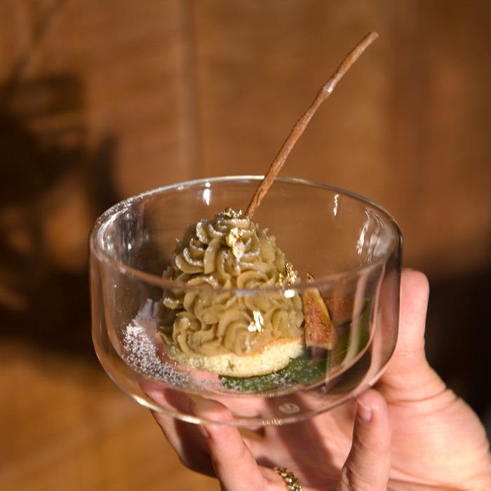 Hand holding a small glass bowl with a dessert resembling a mountain, with a blurred background.