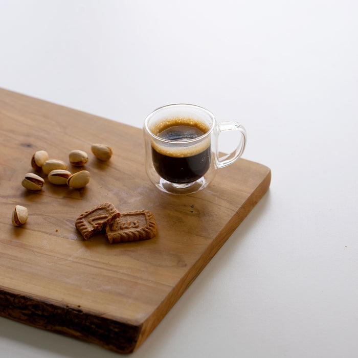 Thermic Sublime Espresso Double Wall Glass filled with espresso and standing on a wooden bard decorated with Lotus cookie and Pistachios