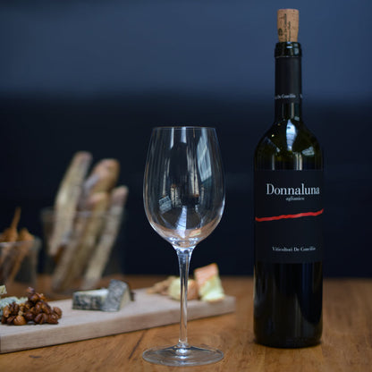 Empty Vinoteque Red Wine Glass standing next to a red wine bottle on a wooden table, behind is a blurred cheese board and a dark background
