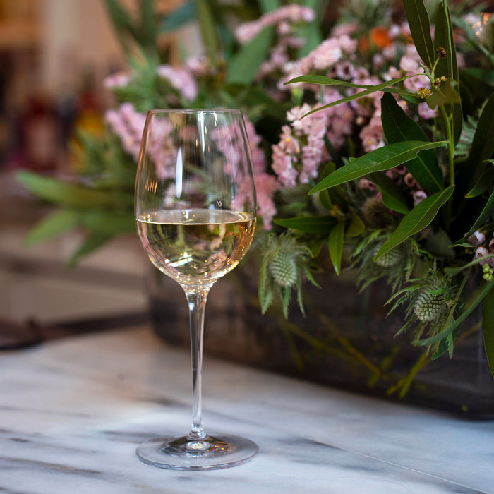 Vinoteque Red Wine Glass filled with white wine standing on a marble surface and a bouquet of pink flowers and greenery in the background