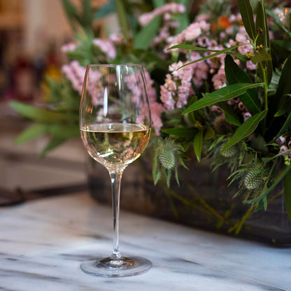 Vinoteque Red Wine Glass filled with white wine standing on a marble surface and a bouquet of pink flowers and greenery in the background