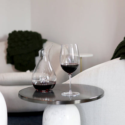 Magnifico decanter filled with red wine standing next to a Vinoteque Riserva wine glass filled with red wine on a silver side table, white chairs are in the background