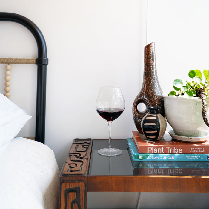 Vinoteque Robusto Red Wine Glass filled with red wine standing on a dark night stand with colorful vases, books and a plant in the background