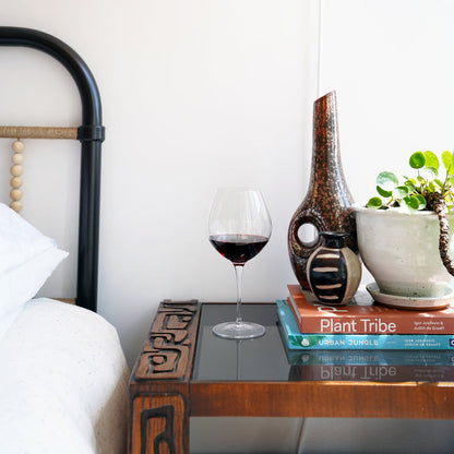 Vinoteque Robusto Red Wine Glass filled with red wine standing on a dark night stand with colorful vases, books and a plant in the background